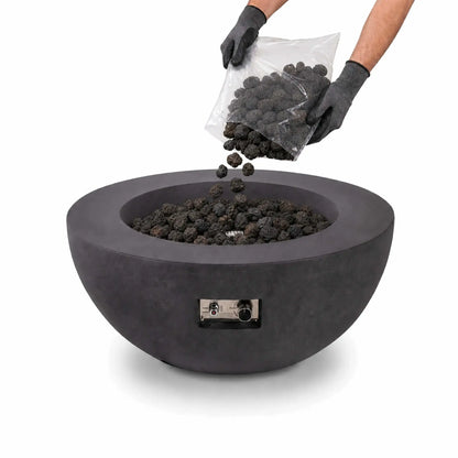 Flame Havens small 1/2-inch standard lava rock being poured from a clear bag into a round charcoal-gray fire pit. A person wearing protective work gloves tips the bag as porous black stones fall into the burner bowl, which is already filled with lava rock. The fire pit’s front-mounted ignition controls are visible against the clean white background.