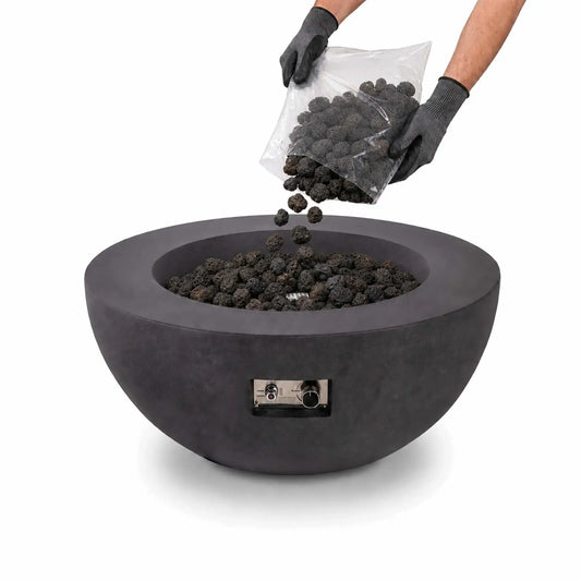 Flame Havens small 1/2-inch standard lava rock being poured from a clear bag into a round charcoal-gray fire pit. A person wearing protective work gloves tips the bag as porous black stones fall into the burner bowl, which is already filled with lava rock. The fire pit’s front-mounted ignition controls are visible against the clean white background.