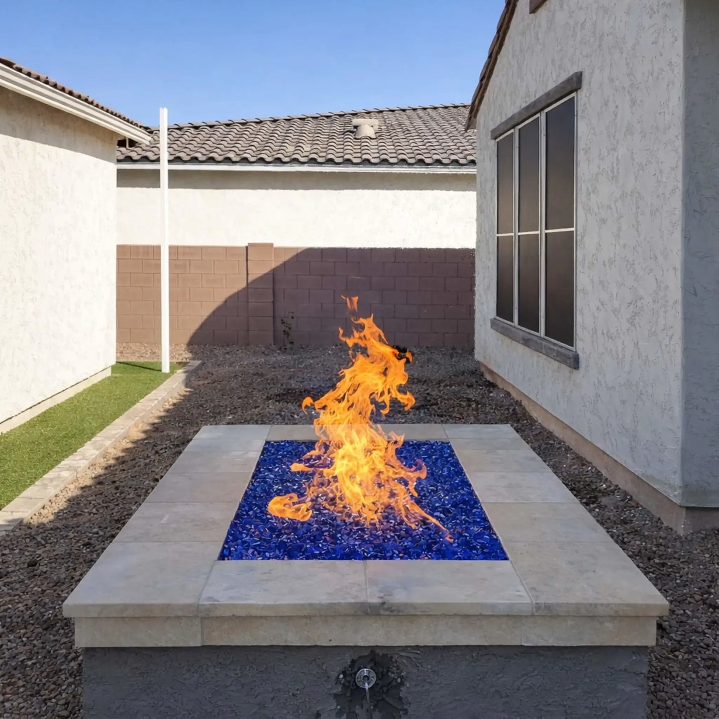 Compact square fire pit with vivid blue fire glass and strong vertical flames, centered in a simple backyard with stucco walls and gravel landscaping. The clean design contrasts with the bright flames, showcasing a modern gas fire feature in a residential outdoor space.