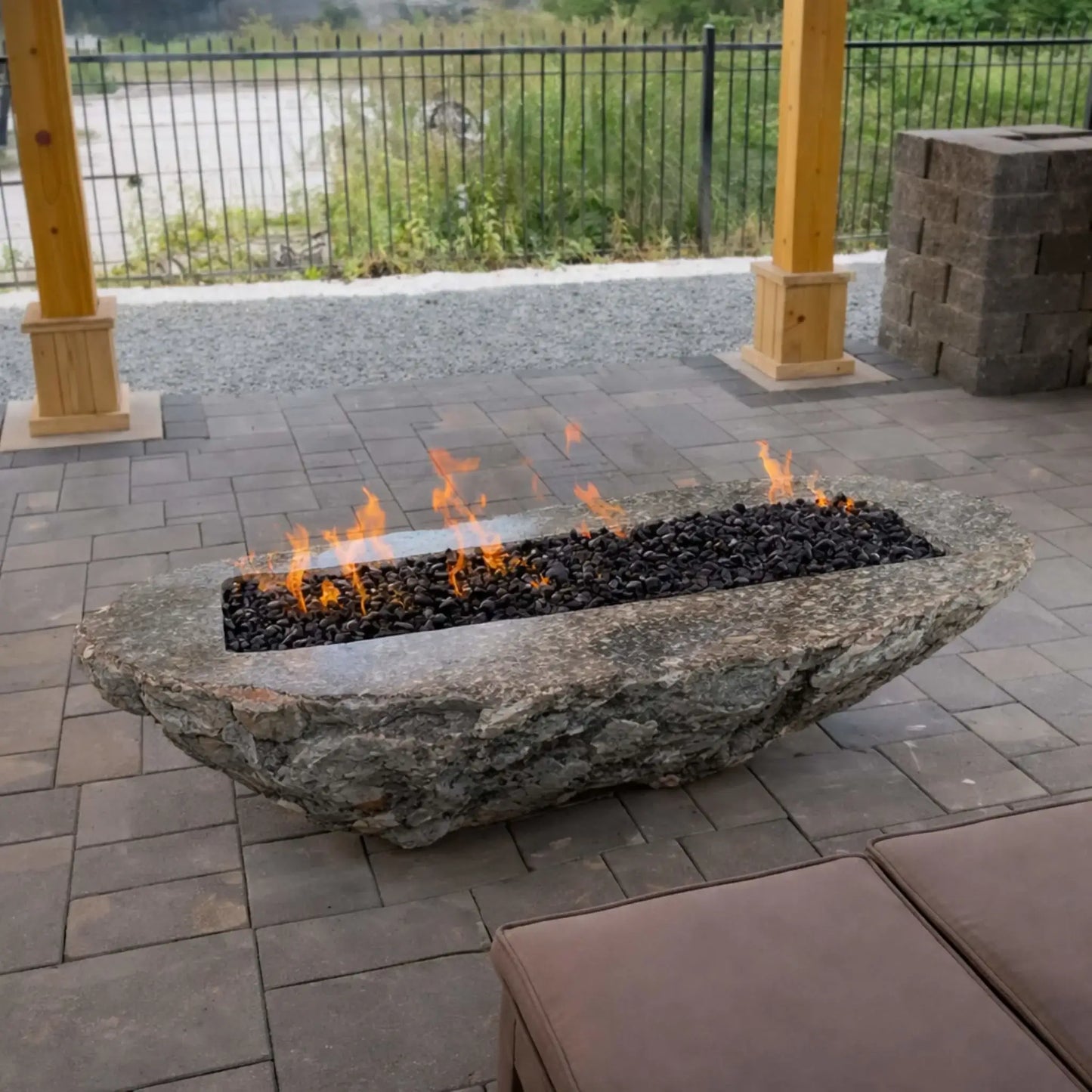 Unique natural stone fire feature carved into a long, rugged boulder, filled with black lava rock and a linear gas flame. Installed on a paved patio beneath a wooden pergola, the design blends organic stone textures with modern fire pit functionality.