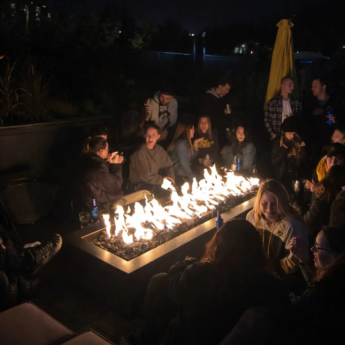 Large group of friends gathered at night around a long rectangular fire pit filled with fire glass, with bright flames illuminating faces and drinks. The scene captures a lively outdoor social setting with warm lighting, conversation, and a cozy fire centerpiece.