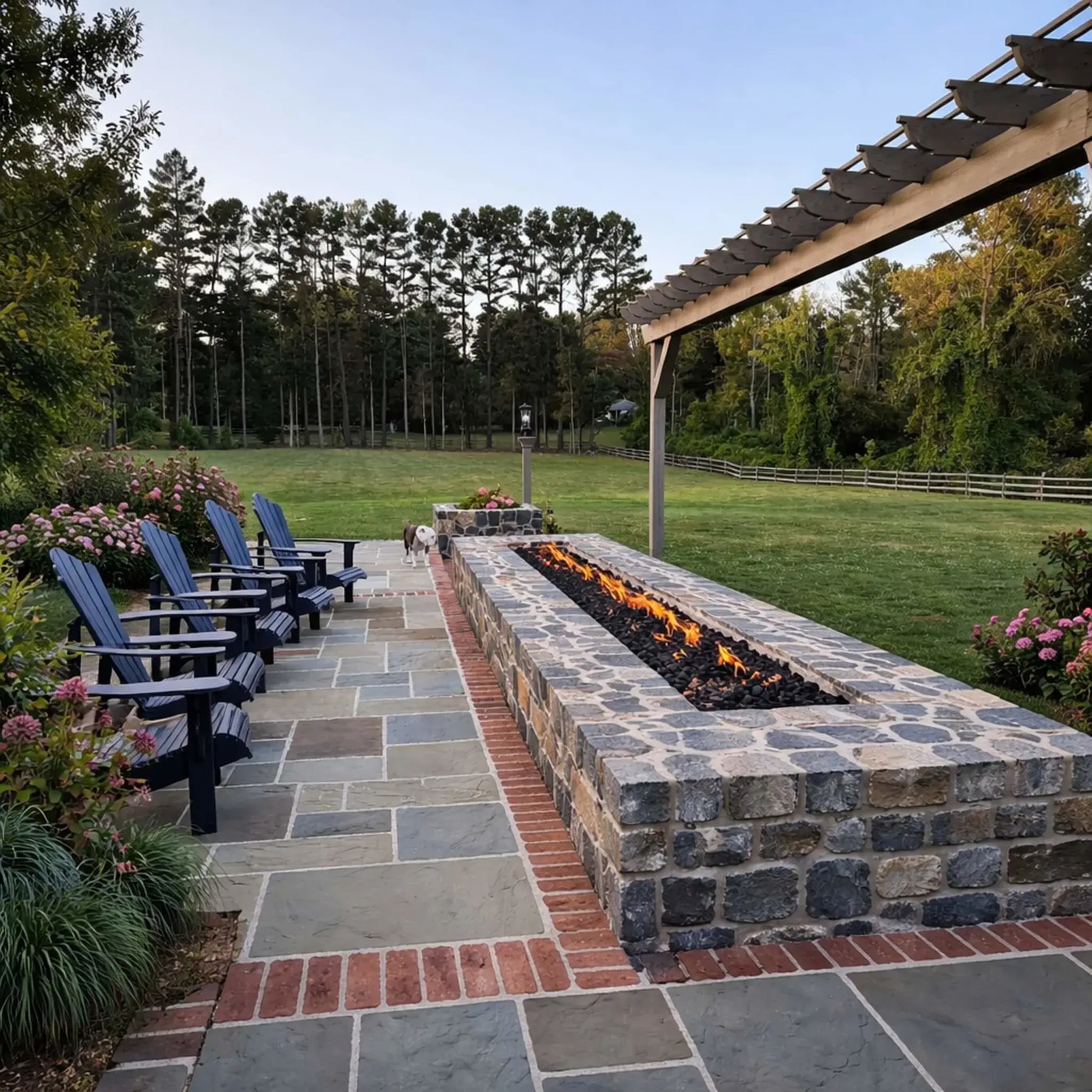 Elegant stone fire pit integrated into a landscaped patio with a pergola overhead and Adirondack chairs lined along the side. A long linear flame runs across the fire feature, set against a wide open lawn and tree line, creating a serene countryside outdoor living space.