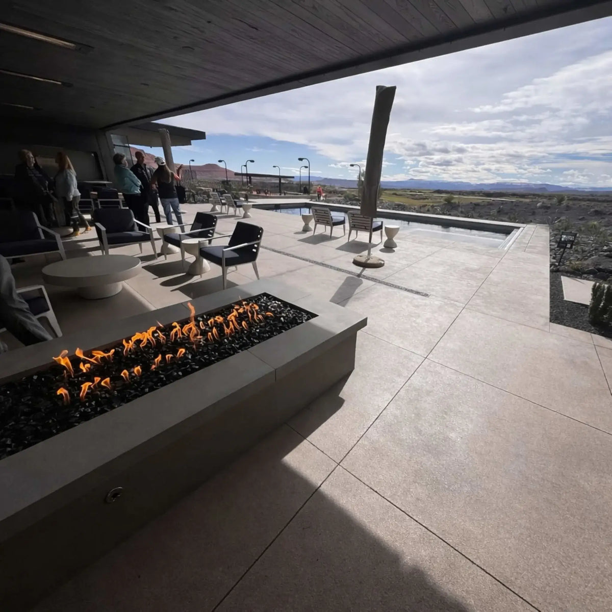 Contemporary covered patio overlooking a scenic desert or mountain landscape, featuring a rectangular gas fire pit filled with black fire glass and a steady linear flame. Modern outdoor furniture and a pool area complete the high-end outdoor entertaining environment.
