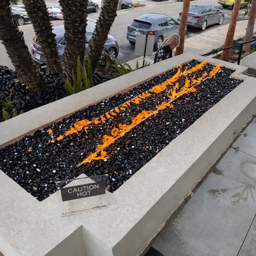 Linear gas fire pit installed along a modern walkway near palm trees and parked cars, filled with black fire glass and producing clean, consistent flames. A small “Caution Hot” sign sits on the edge, emphasizing safety in this sleek commercial or urban outdoor setting.