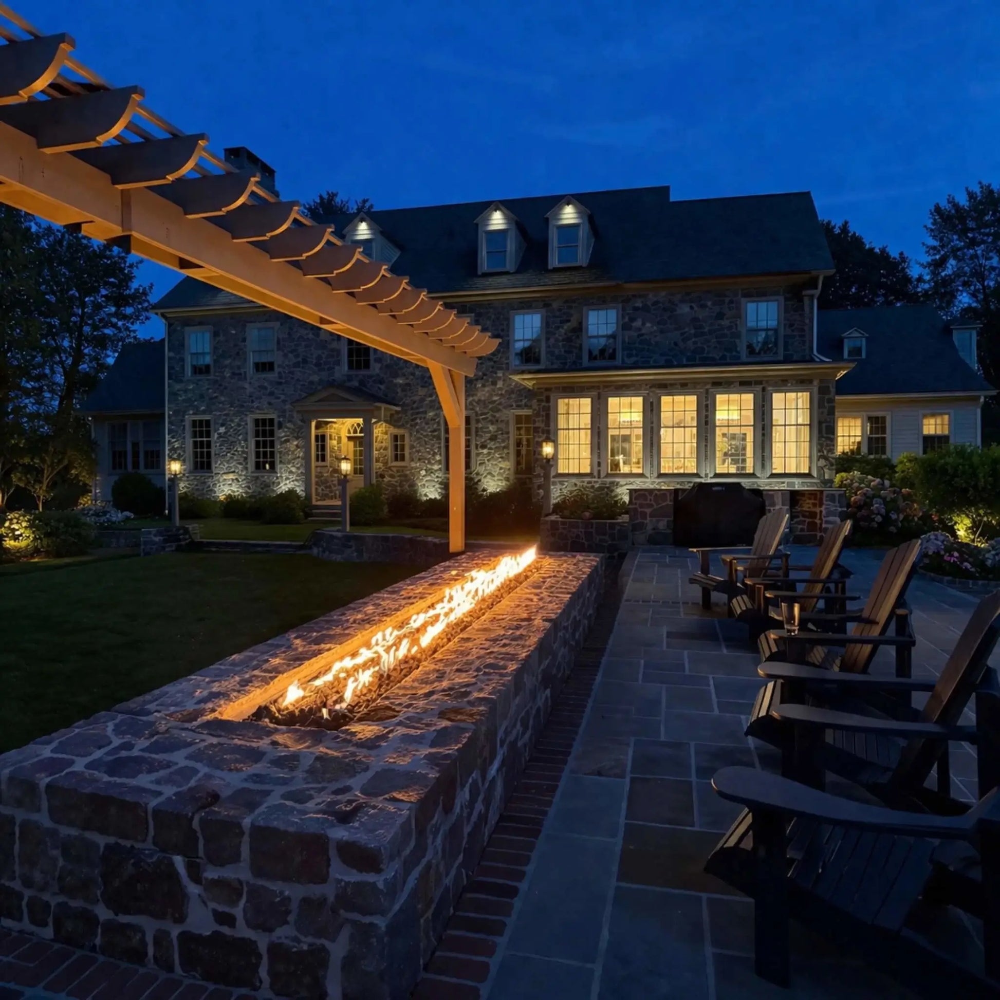 Evening backyard scene with a long stone fire feature beneath a pergola, glowing with a continuous linear flame. A large, well-lit home sits in the background, while patio chairs line the space, creating a warm and inviting outdoor gathering area at dusk.