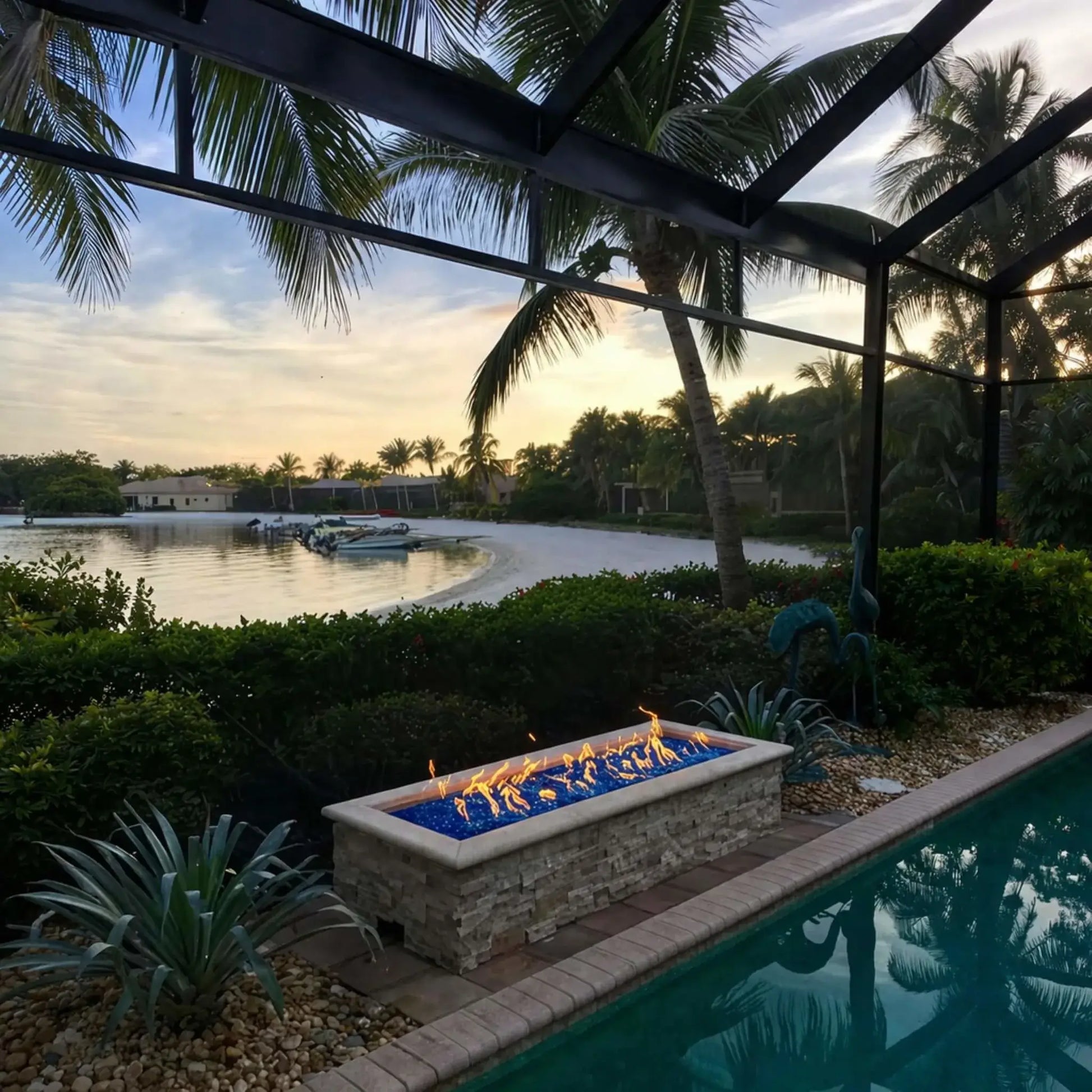 Modern rectangular fire pit filled with vibrant blue fire glass burning along a waterfront patio at sunset, surrounded by palm trees and a screened enclosure. The flames stretch across the burner, complementing the tropical landscaping and calm water views for a luxury outdoor living space.