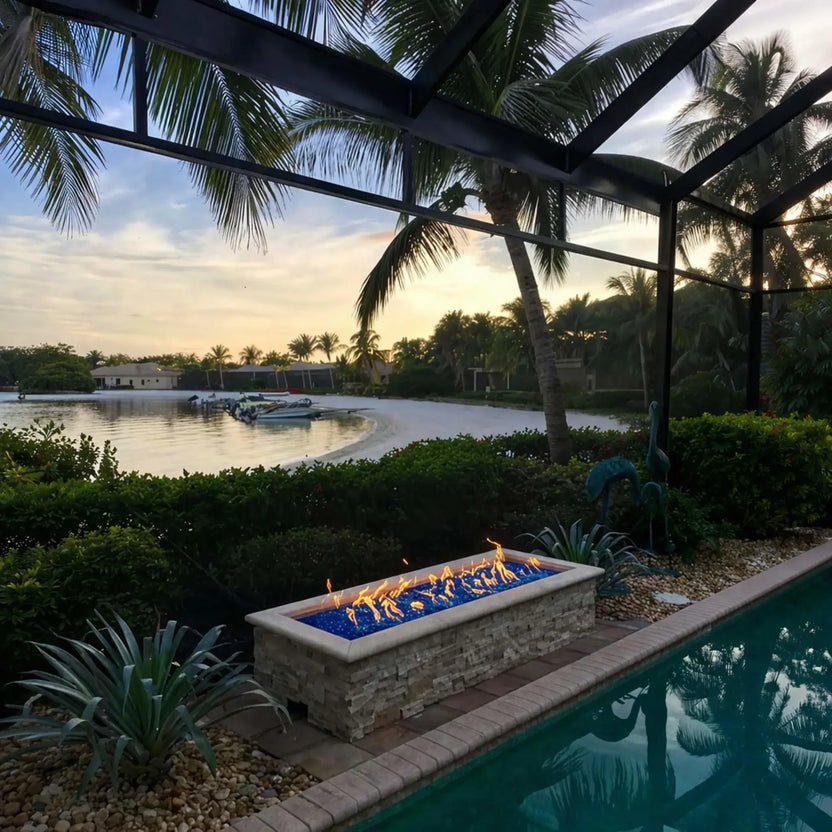 Modern rectangular fire pit filled with vibrant blue fire glass burning along a waterfront patio at sunset, surrounded by palm trees and a screened enclosure. The flames stretch across the burner, complementing the tropical landscaping and calm water views for a luxury outdoor living space.