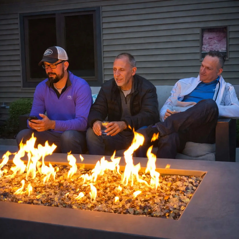 Three men relaxing on outdoor patio seating around a long rectangular gas fire pit filled with reflective fire glass. The flames flicker evenly across the burner, casting warm light as the group enjoys drinks and conversation in a casual backyard evening setting.