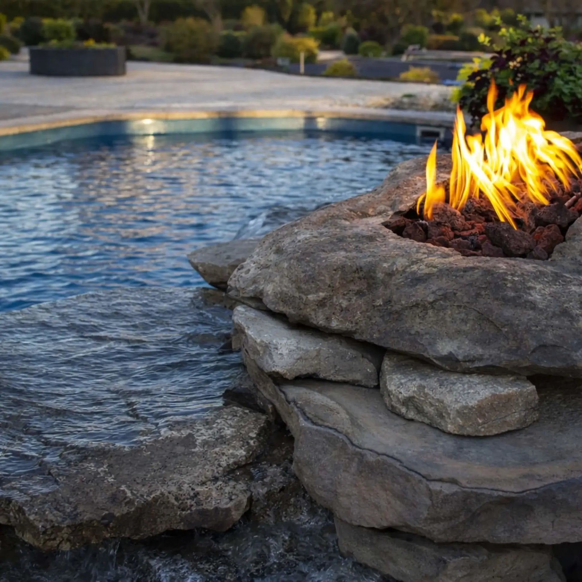Natural stone fire feature integrated beside a pool, with flames rising from lava rock set into a rugged rock formation. Water gently cascades nearby, creating a striking combination of fire and water elements in a landscaped backyard pool environment.