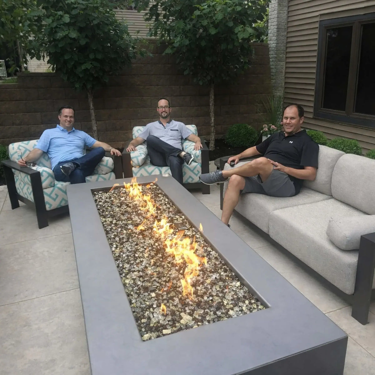 Spacious backyard patio featuring a large rectangular fire pit table filled with decorative fire glass and a linear flame pattern. Three men sit comfortably on cushioned outdoor furniture, enjoying a relaxed atmosphere with trees and privacy walls in the background.