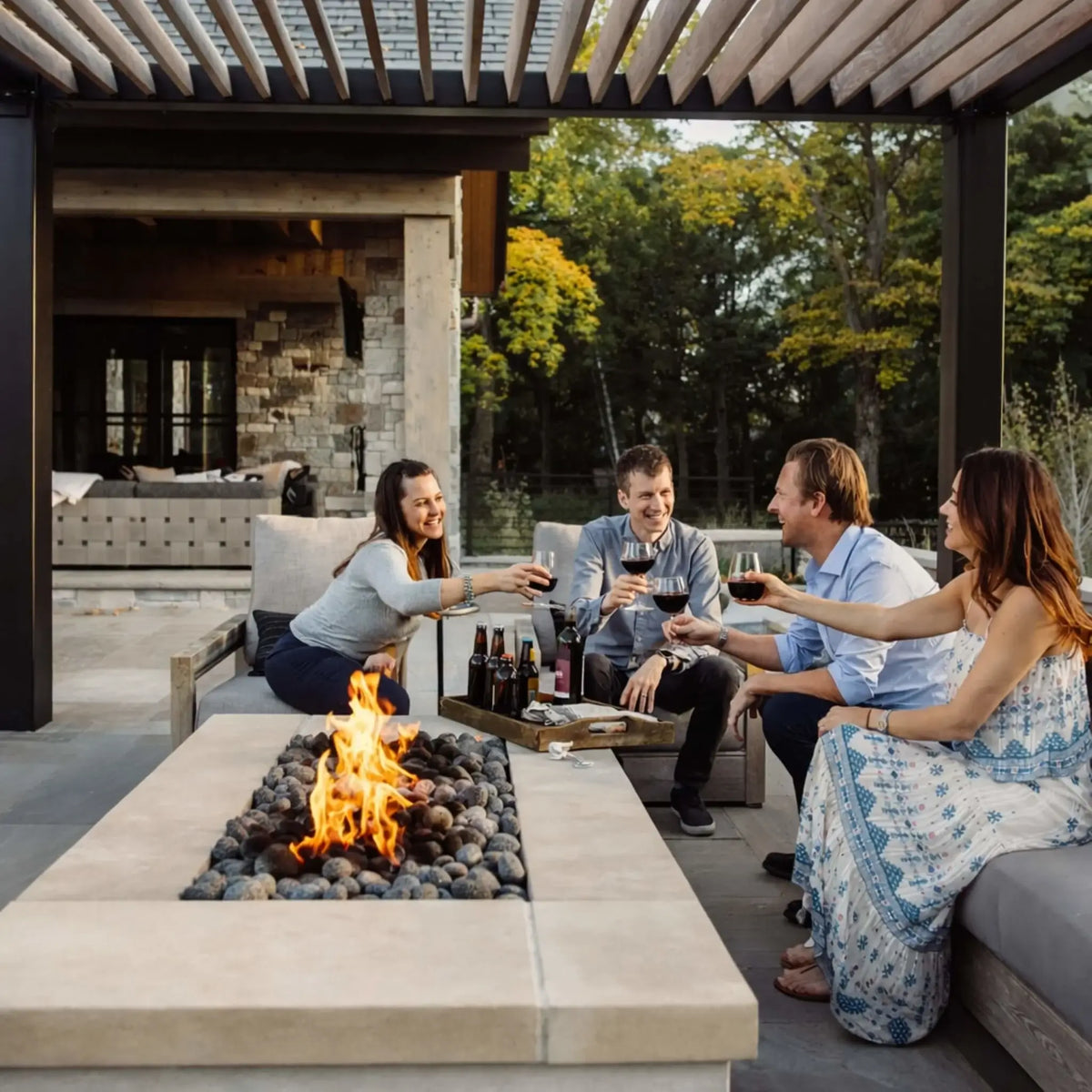 Group of friends gathered around a modern outdoor fire pit table under a pergola, raising glasses in a relaxed social setting. The linear gas burner produces steady flames over smooth stones, complementing the upscale patio design and evening outdoor entertaining atmosphere.