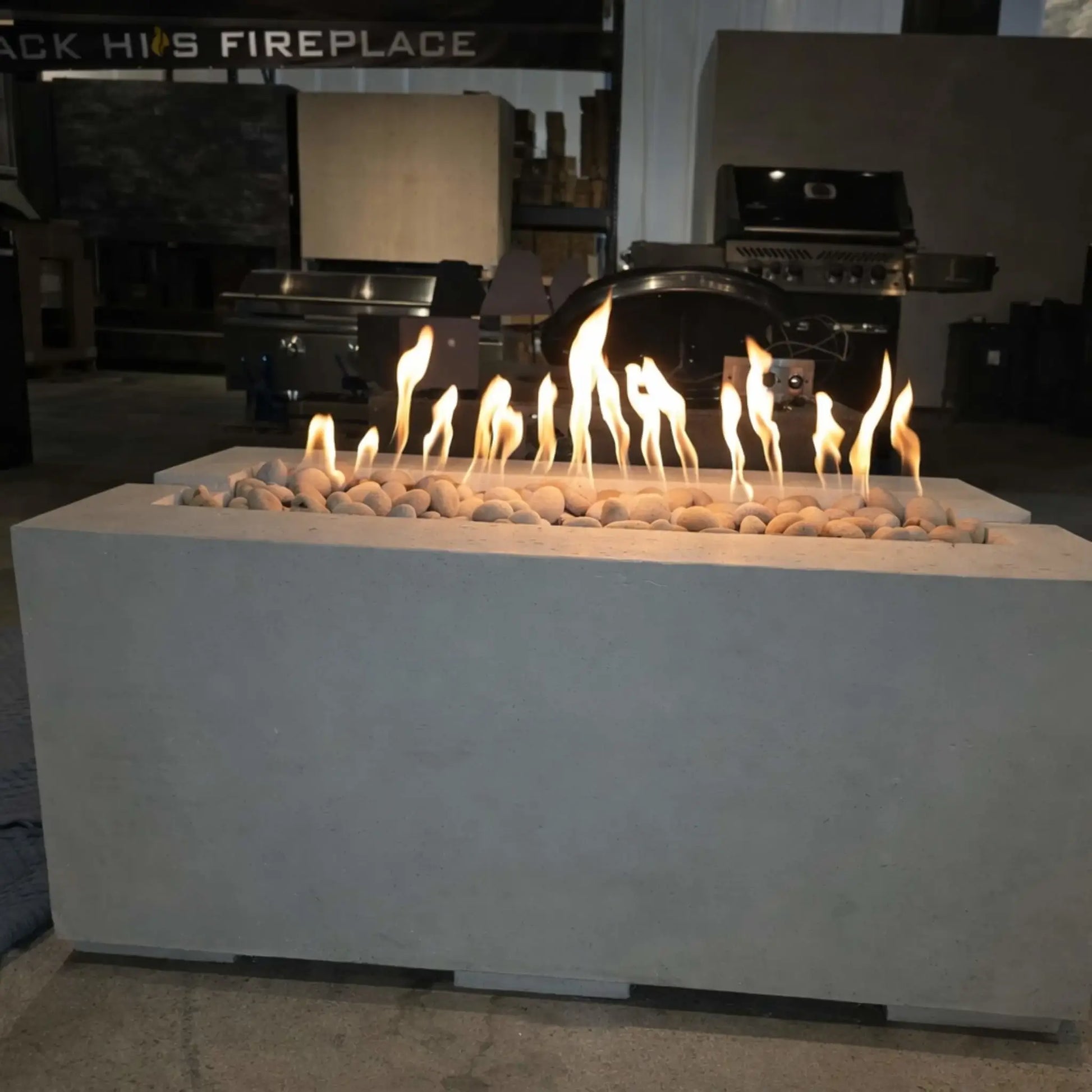 A modern rectangular concrete fire pit table displayed indoors, featuring a linear gas burner filled with smooth white river stones. Bright, even flames rise across the center, while stainless steel grills and outdoor kitchen equipment in the background suggest a showroom setting for premium fire features.