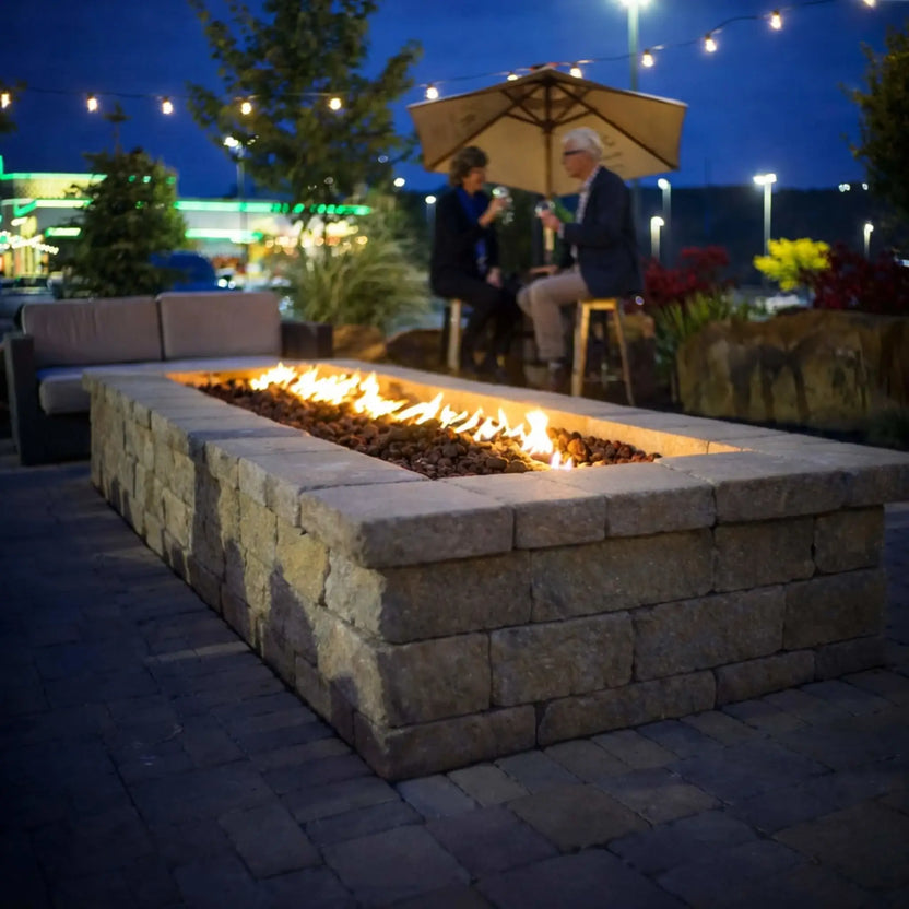 A cozy outdoor patio at night with a large rectangular stone fire pit table glowing warmly. A couple sits nearby under a patio umbrella with string lights overhead, enjoying drinks while the linear flame stretches across lava rock media, creating a welcoming, social atmosphere.