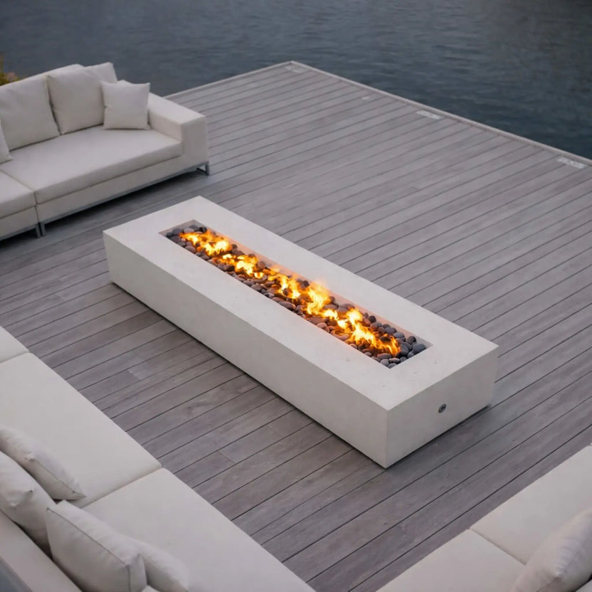 A sleek waterfront deck setup with a long, low-profile concrete fire pit centered between plush outdoor sofas. The linear flame burns steadily over dark fire glass, complementing the calm water backdrop and minimalist outdoor design for a serene, modern lounging space.