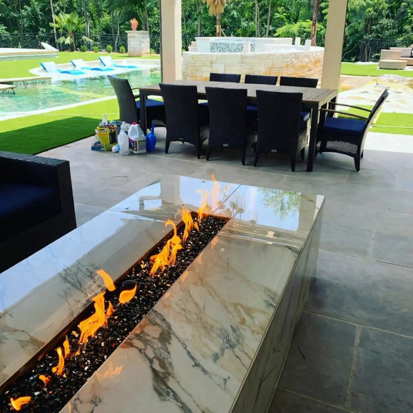 A covered backyard patio featuring a polished stone fire table with a linear burner filled with black fire glass. Flames flicker brightly in the foreground, while a dining table, pool, and landscaped yard create a luxurious outdoor entertainment area.