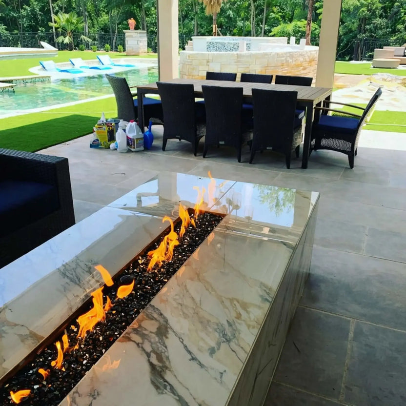 A covered backyard patio featuring a polished stone fire table with a linear burner filled with black fire glass. Flames flicker brightly in the foreground, while a dining table, pool, and landscaped yard create a luxurious outdoor entertainment area.