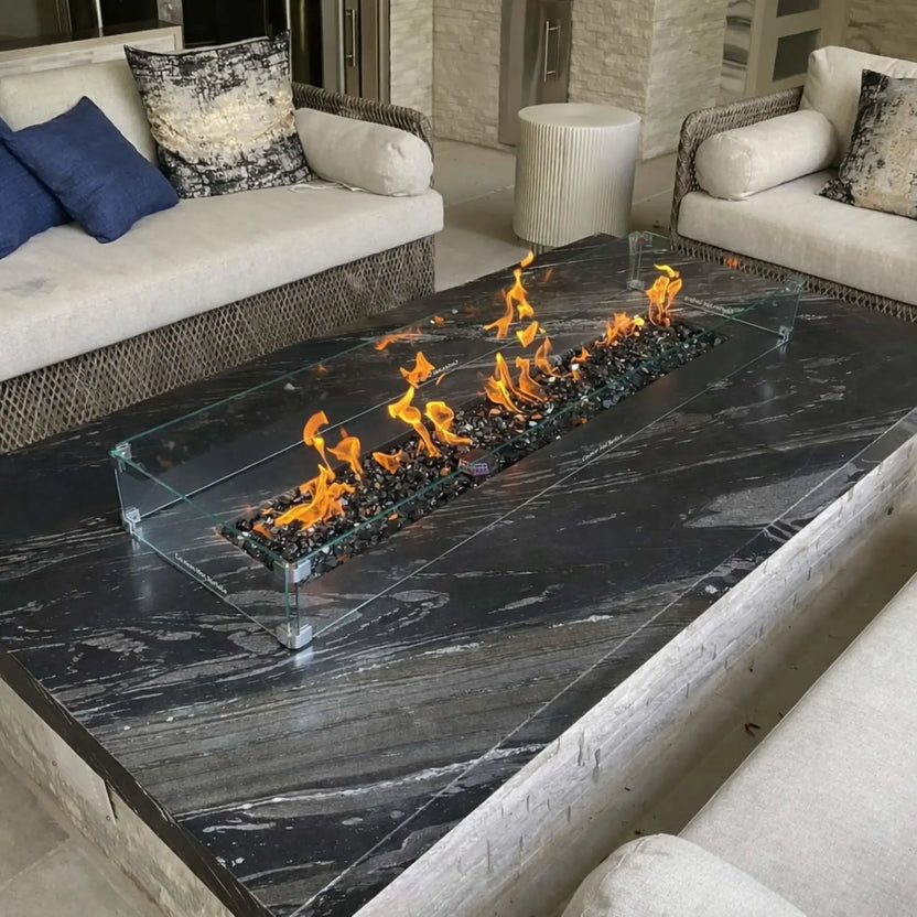 A contemporary outdoor seating area with a black marble fire table and a linear gas burner protected by a clear glass wind guard. The flames dance across dark fire media, surrounded by cushioned wicker seating and textured pillows for a refined, upscale patio setting.