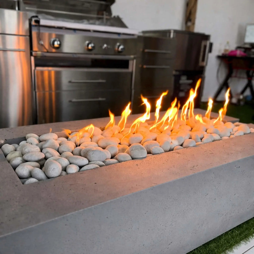 A close-up of a rectangular concrete fire pit filled with smooth white stones, showcasing a clean-burning linear flame. Set in front of stainless steel outdoor appliances, the image highlights the burner’s even flame pattern and modern design appeal.