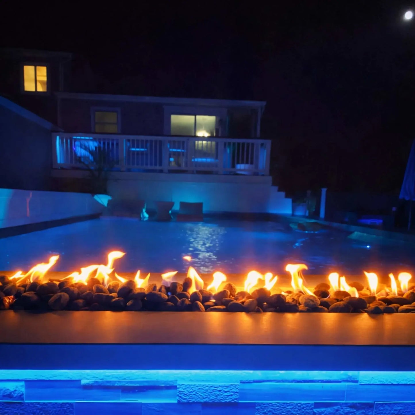 A nighttime backyard pool scene illuminated with blue accent lighting and a long linear fire feature in the foreground. Flames flicker over dark stones, reflecting across the water while a raised deck and house glow softly in the background.