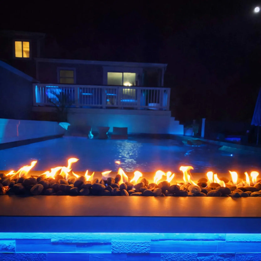 A nighttime backyard pool scene illuminated with blue accent lighting and a long linear fire feature in the foreground. Flames flicker over dark stones, reflecting across the water while a raised deck and house glow softly in the background.