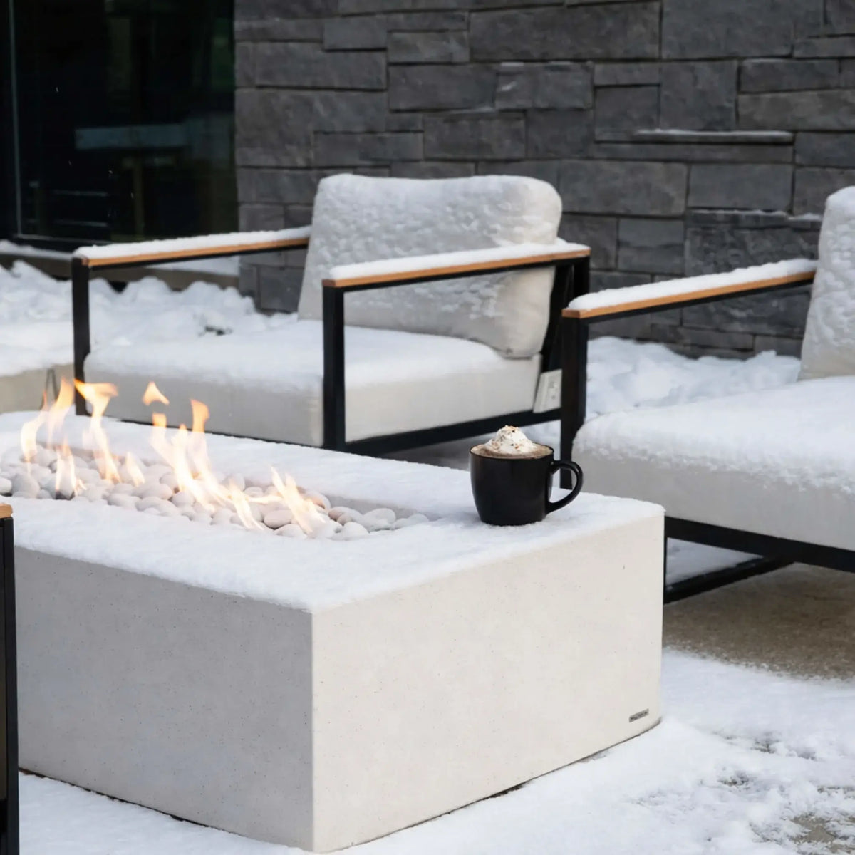 A winter patio setting with a rectangular concrete fire pit surrounded by snow-covered outdoor chairs. A warm linear flame burns through white stones, contrasting against the cold environment, with a mug of hot cocoa placed nearby for a cozy seasonal touch.
