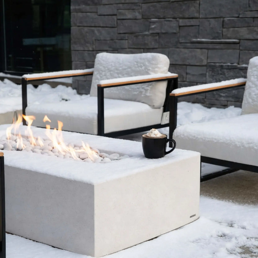 A winter patio setting with a rectangular concrete fire pit surrounded by snow-covered outdoor chairs. A warm linear flame burns through white stones, contrasting against the cold environment, with a mug of hot cocoa placed nearby for a cozy seasonal touch.