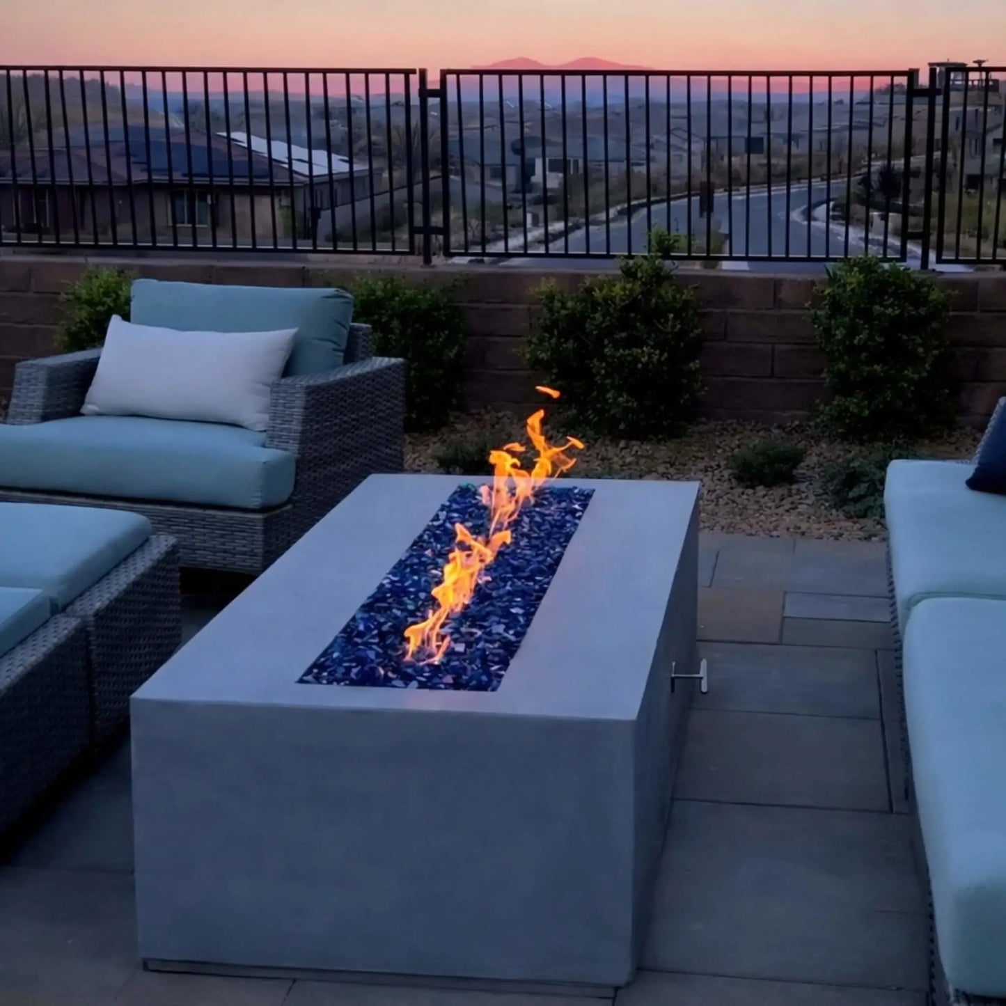 A modern backyard patio at sunset featuring a square fire table with a linear flame running through blue fire glass. Surrounded by cushioned seating and a metal fence overlooking rolling hills, the fire feature creates a relaxing ambiance as the sky glows in soft evening tones.