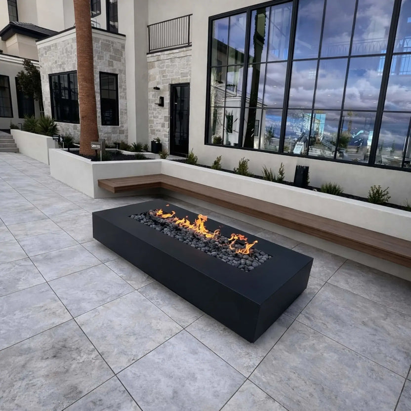 A modern luxury courtyard featuring a sleek black rectangular linear fire pit filled with dark lava rock. Set against a contemporary home with large floor-to-ceiling windows, stone accents, and built-in bench seating, the clean flame line adds warmth and contrast to the minimalist outdoor design.