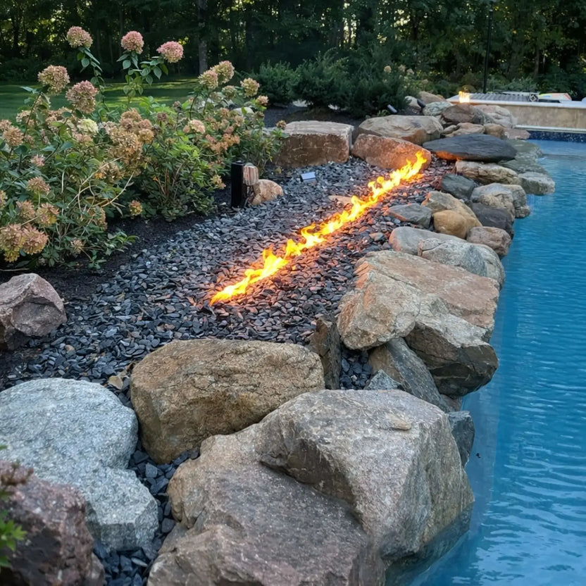 A natural-style poolside landscape with a linear fire burner installed among dark gravel and large boulders. The flame runs in a clean line beside the pool edge, adding warmth and contrast to the surrounding greenery, shrubs, and soft hydrangea blooms in a peaceful backyard setting.