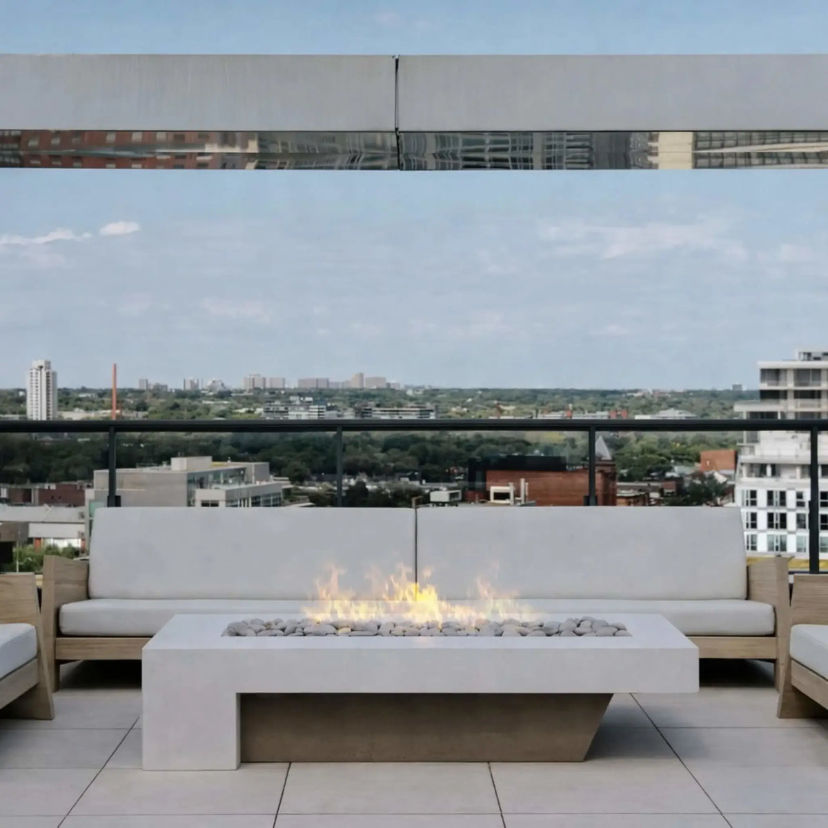 A rooftop patio with a white linear fire pit table centered between cushioned outdoor sofas. The soft flames rise over white stones, while a glass railing reveals a city skyline view in the distance, creating a calm, elevated urban lounge setting.