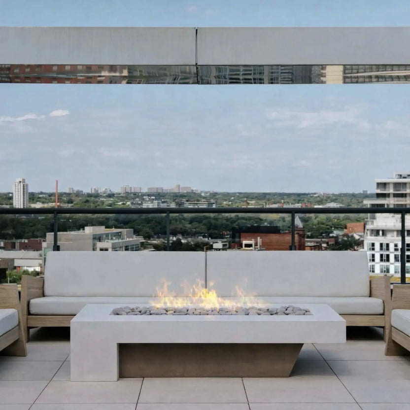 A rooftop patio with a white linear fire pit table centered between cushioned outdoor sofas. The soft flames rise over white stones, while a glass railing reveals a city skyline view in the distance, creating a calm, elevated urban lounge setting.