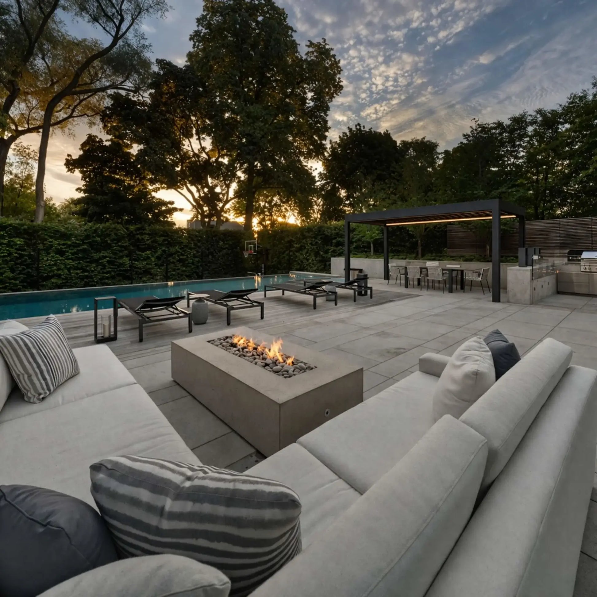 A spacious backyard patio at sunset featuring a rectangular concrete fire pit surrounded by plush seating. Beyond the fire feature, a pool, lounge chairs, and a covered outdoor kitchen area sit under tall trees, creating a relaxed, resort-style outdoor living space.