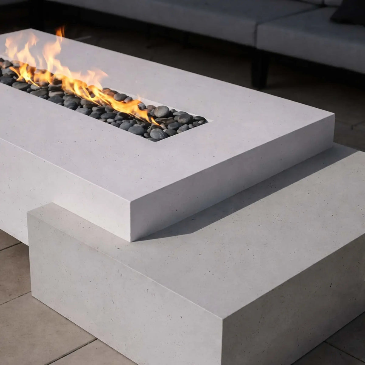 A close-up of a modern white concrete fire table with a recessed linear burner filled with smooth gray stones. The flames run cleanly across the center, highlighting the crisp edges and layered design of the fire pit in a contemporary patio setting.