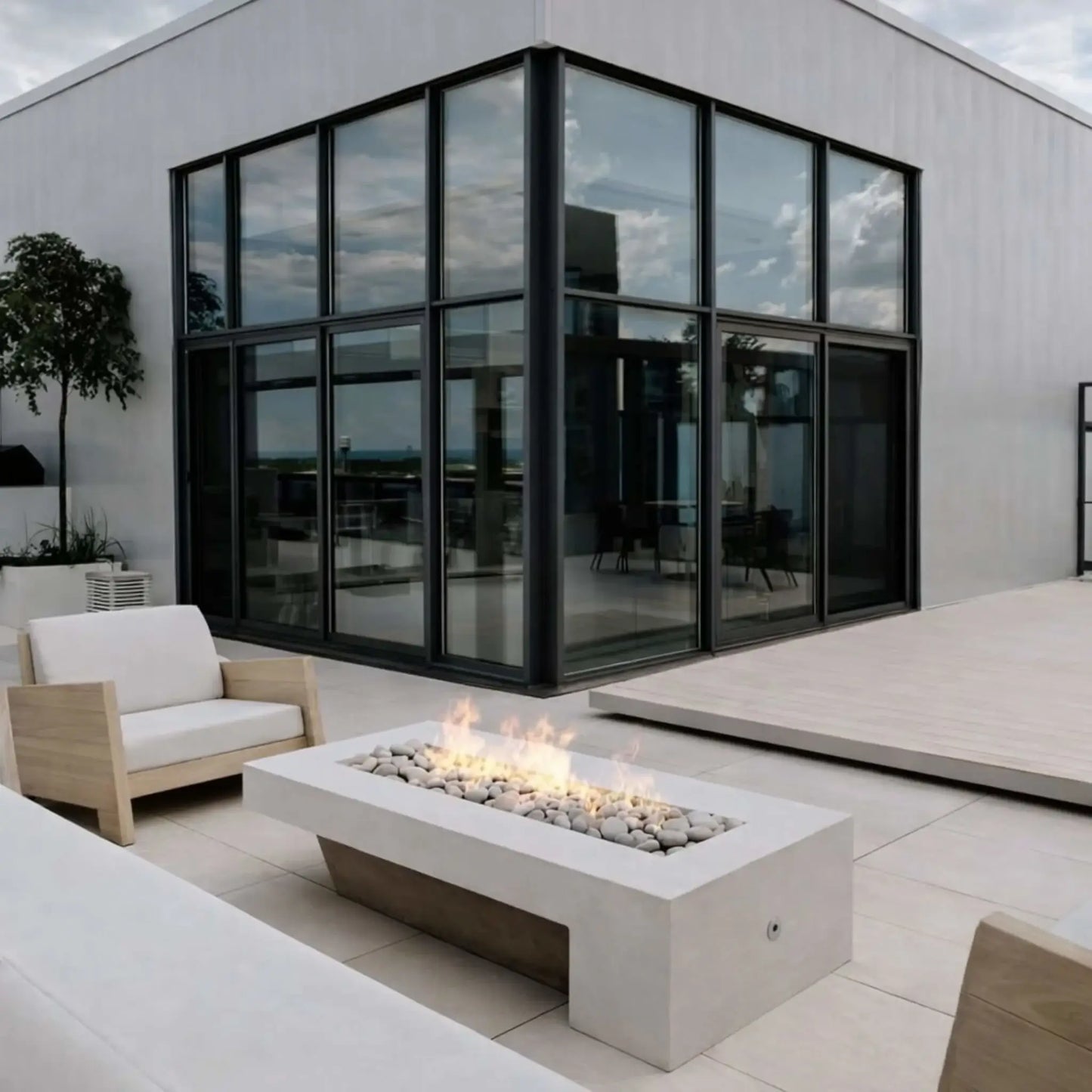 A modern outdoor seating area featuring a low-profile rectangular fire table with white stones and a steady linear flame. Surrounded by light wood-framed chairs and neutral cushions, the setup sits against a sleek gray wall, offering a minimalist and cozy ambiance.