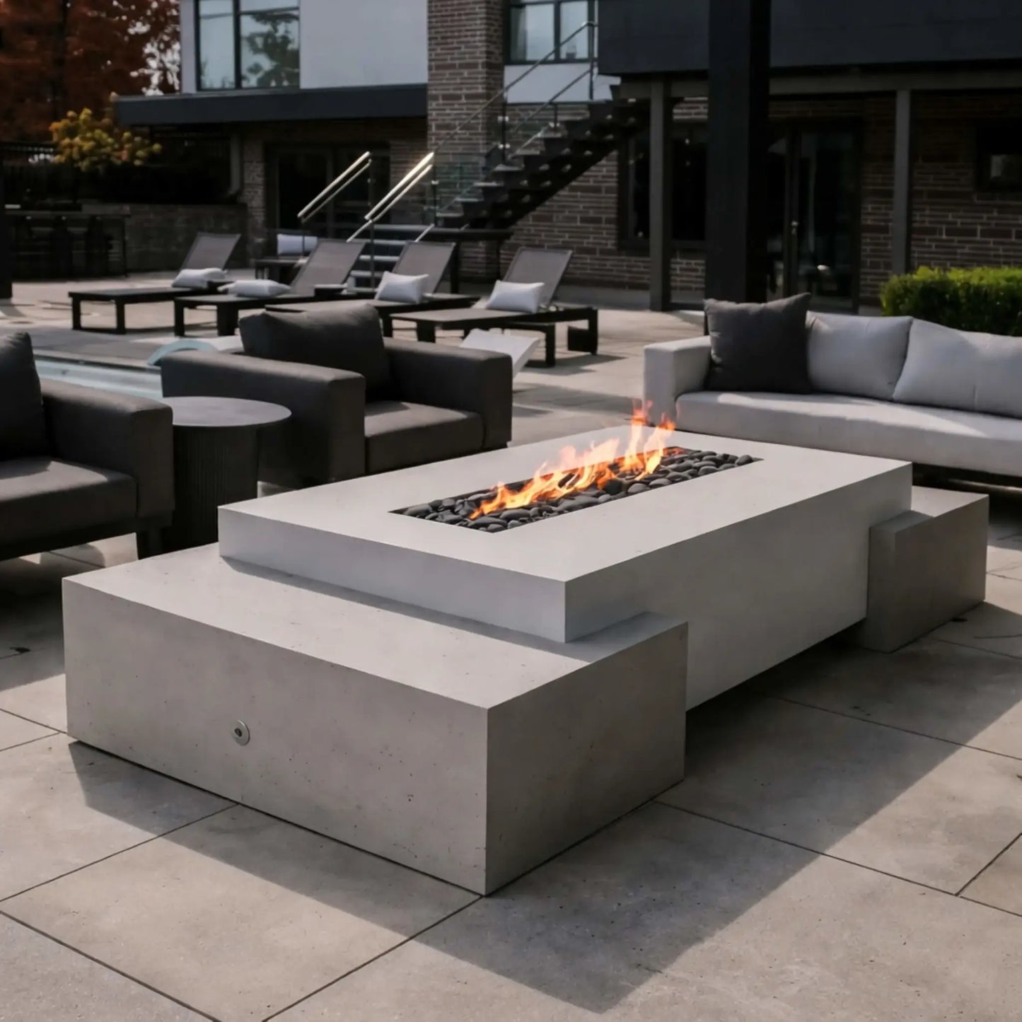 A backyard patio at dusk featuring a stone-built rectangular fire feature with dual linear flames. Positioned near an outdoor dining table and umbrella, the warm firelight enhances the landscaped surroundings and creates an inviting evening gathering space.