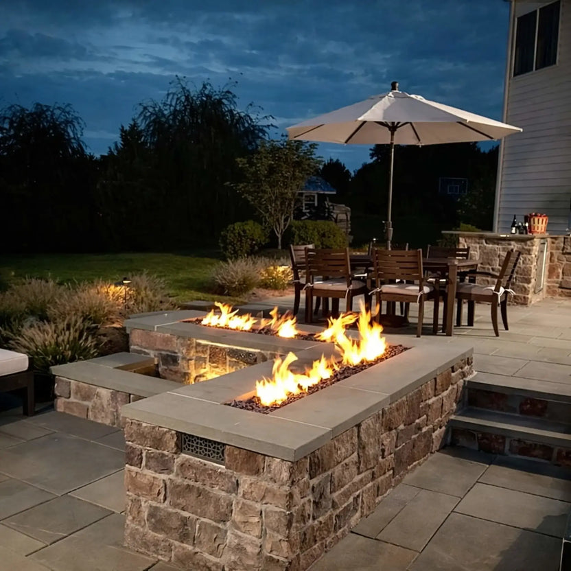 A contemporary deck with a rectangular concrete fire pit surrounded by modern metal-framed chairs. Flames rise above white river stones, while large glass doors reveal a warmly lit interior, blending indoor and outdoor living in a cozy evening setting.