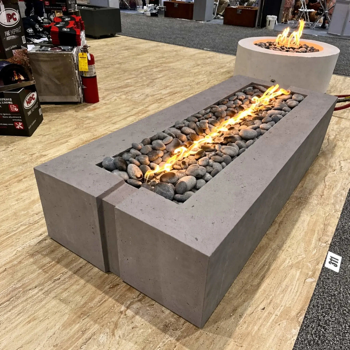 A rectangular concrete gas fire pit on display at an indoor trade show, featuring a Torpedo T linear burner with a continuous flame running through smooth gray river rocks. The unit sits on a wood-look floor with other fire pit displays, fire extinguishers, and product booths visible in the background.
