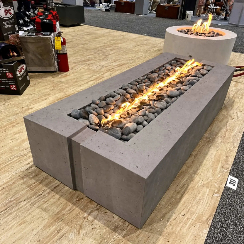 A rectangular concrete gas fire pit on display at an indoor trade show, featuring a Torpedo T linear burner with a continuous flame running through smooth gray river rocks. The unit sits on a wood-look floor with other fire pit displays, fire extinguishers, and product booths visible in the background.