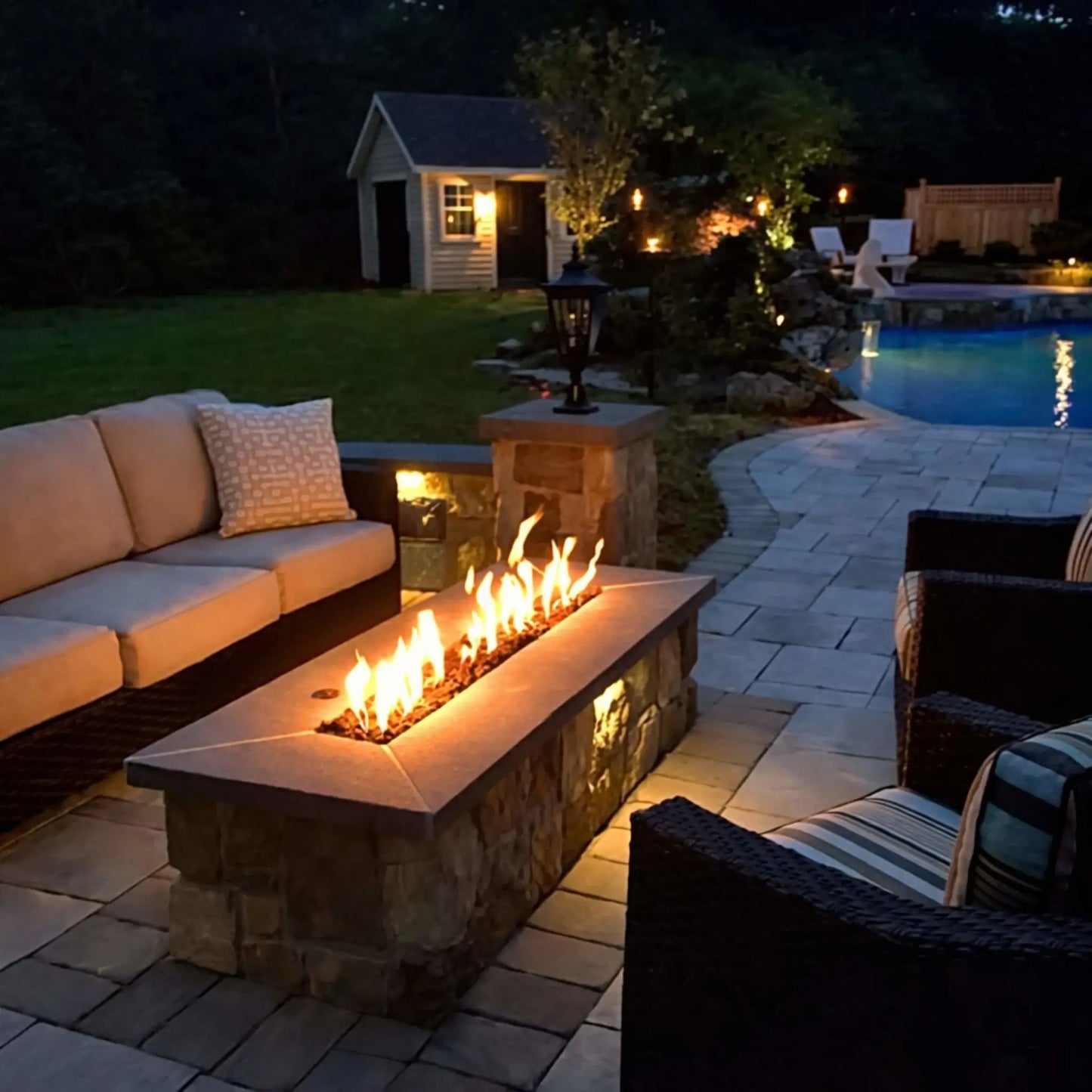 A cozy backyard patio at night featuring a stone-clad rectangular fire pit with a bright linear flame. Surrounded by cushioned outdoor seating, landscape lighting, and a nearby pool with a waterfall feature, the warm glow creates an inviting outdoor living space.