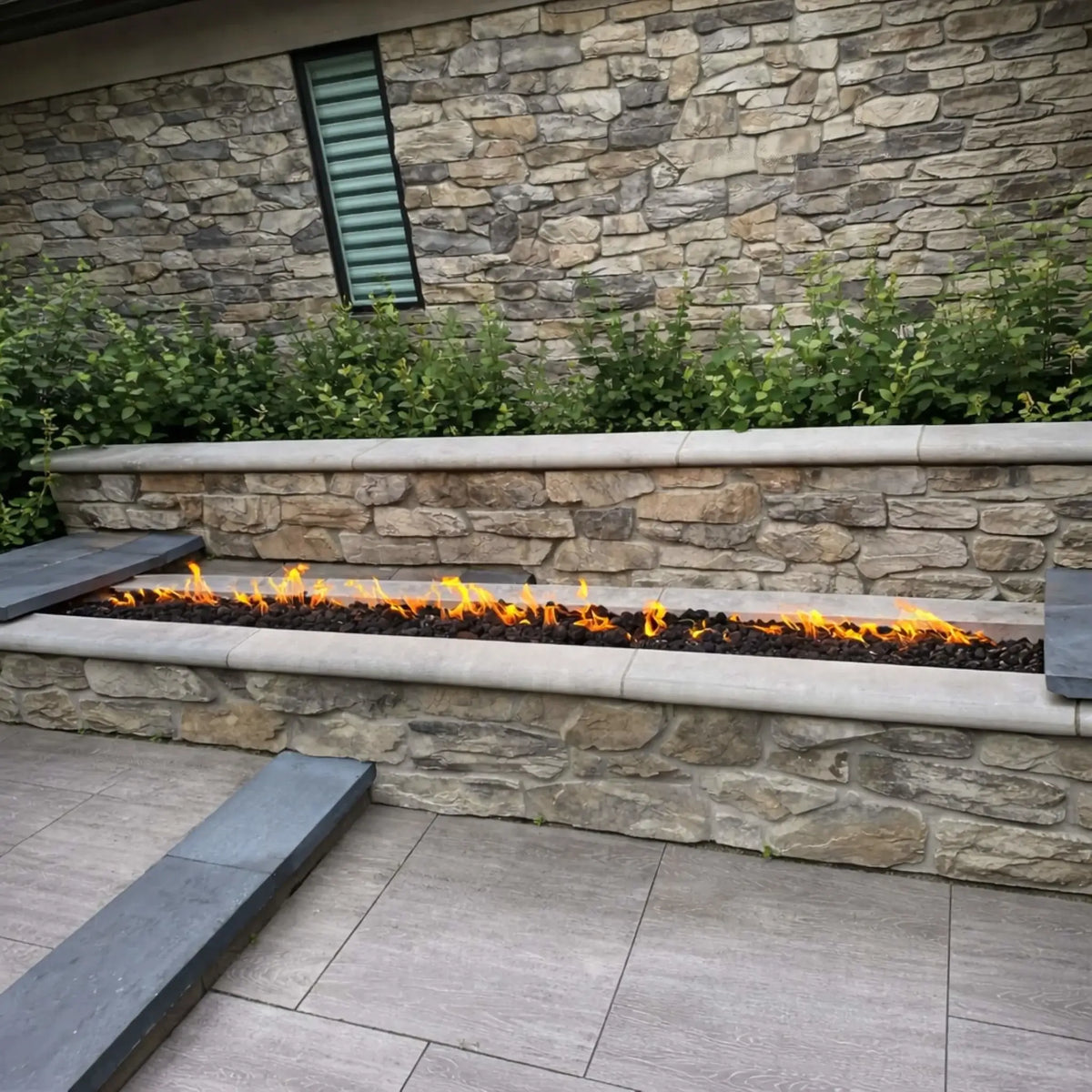 A close-up view of a linear gas fire burner installed in a sleek stone fire table next to a pool. The flames rise evenly from a stainless steel burner, reflecting off the smooth surface while wet spots from recent use add a realistic, lived-in feel to the outdoor space.