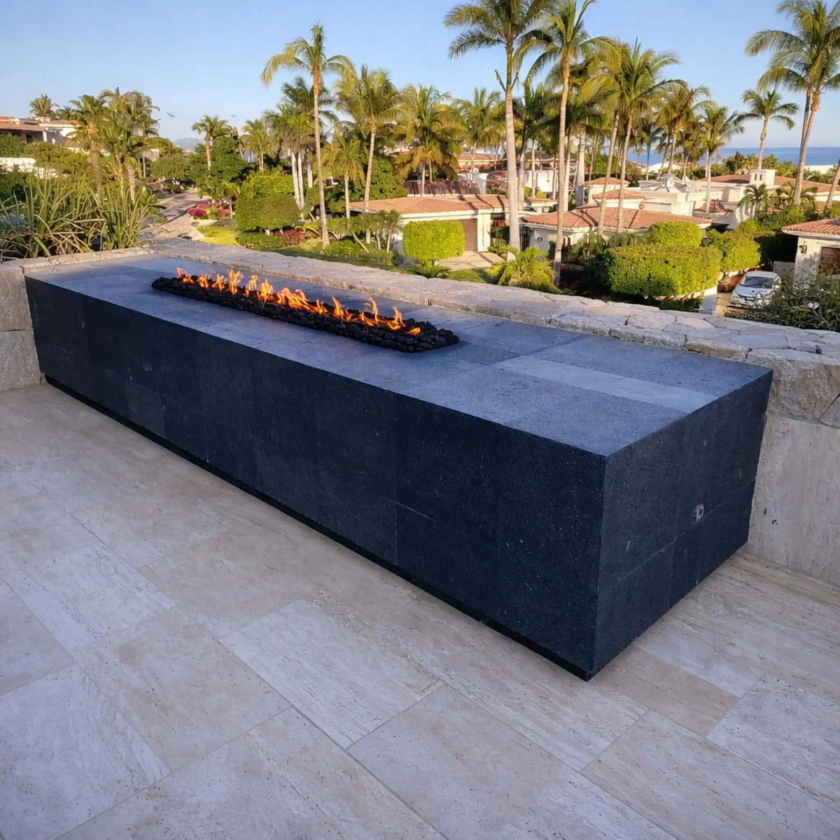 A cozy rooftop or elevated deck space featuring a long linear fire feature with glass wind guards. Two modern woven lounge chairs with blue cushions face the fire, while a lake and sunset view in the distance create a relaxing, upscale outdoor retreat.