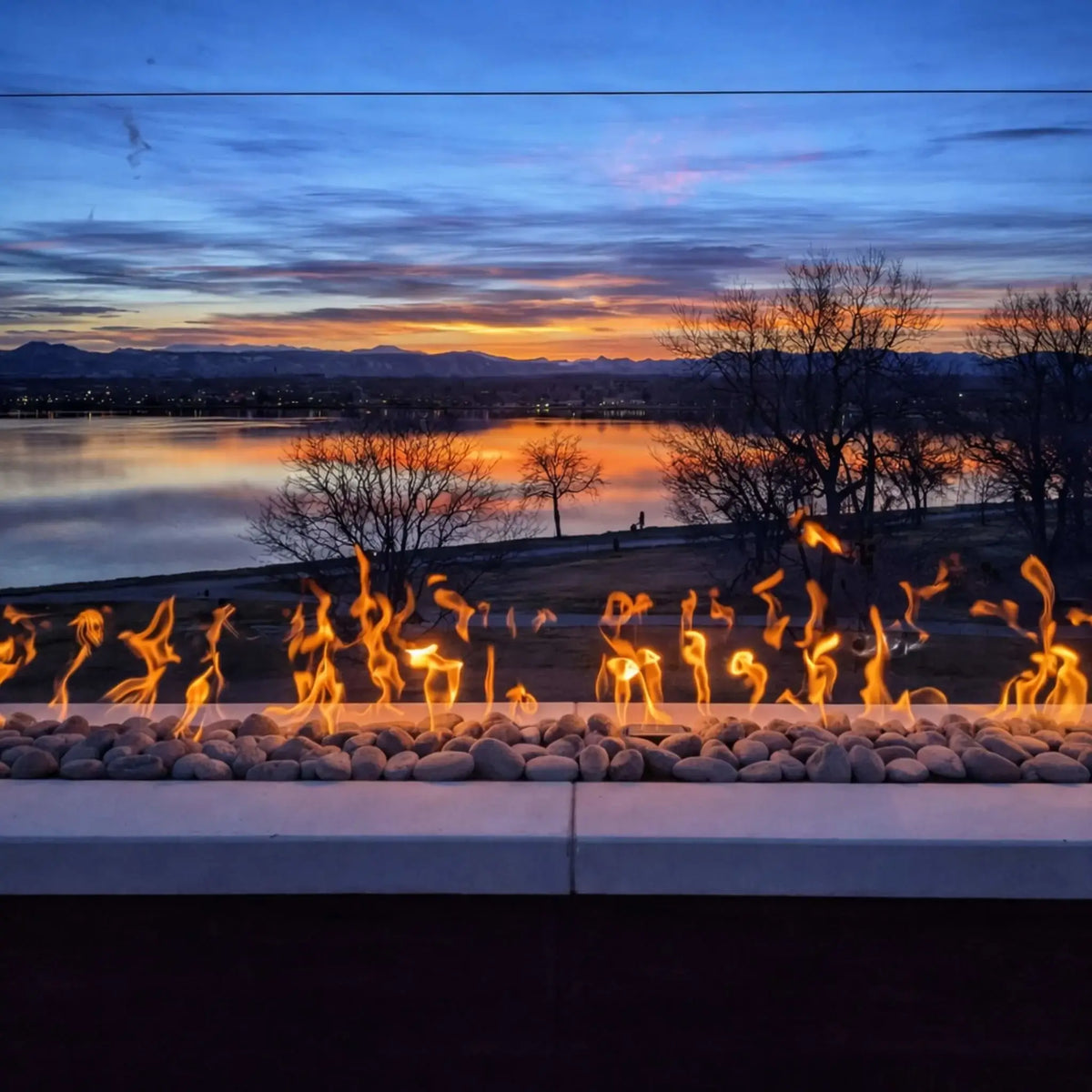 A clean, modern linear fire pit design built into a rectangular structure filled with dark lava rock and accented with small glowing embers. Positioned on a spacious patio near a covered spa, the fire feature adds subtle warmth and elegance to a lakeside outdoor space.