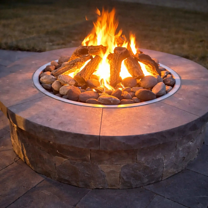 Round stone gas fire pit with Arizona weathered oak logs arranged in a natural campfire stack, surrounded by smooth river rocks. Bright flames rise at dusk, casting a warm glow across the stone patio, highlighting the realistic bark texture and split log detail for a cozy, inviting outdoor atmosphere.