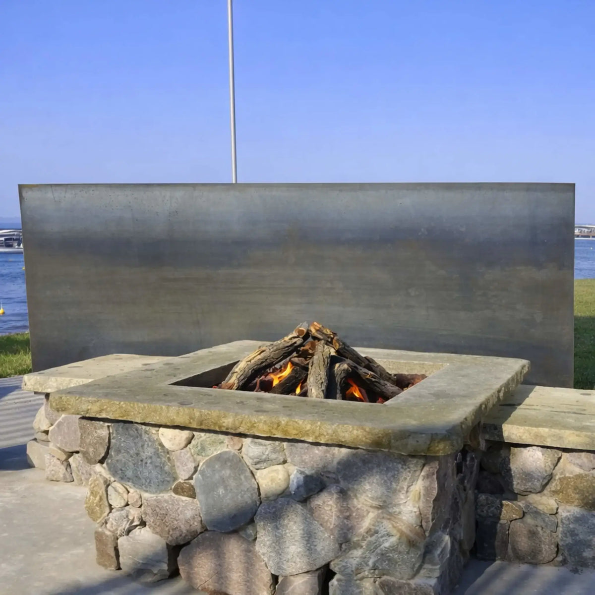 Square stone gas fire pit with weathered oak logs burning in a teepee-style arrangement, set against a modern metal windscreen near a waterfront. Subtle flames flicker between the logs, showcasing a clean, contemporary outdoor design that blends natural stone with sleek architectural elements.