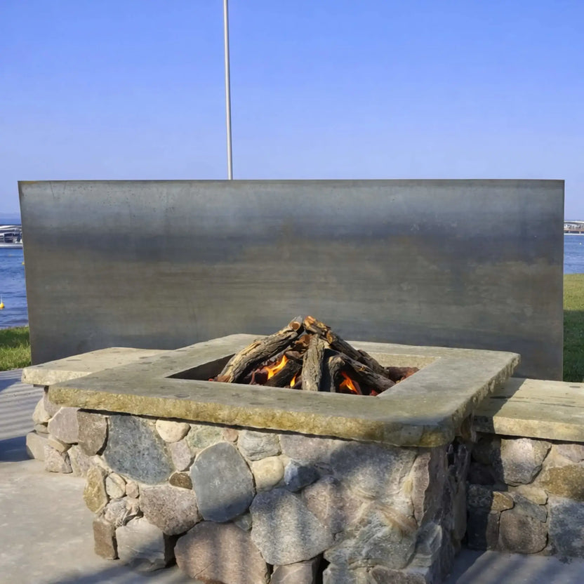 Square stone gas fire pit with weathered oak logs burning in a teepee-style arrangement, set against a modern metal windscreen near a waterfront. Subtle flames flicker between the logs, showcasing a clean, contemporary outdoor design that blends natural stone with sleek architectural elements.