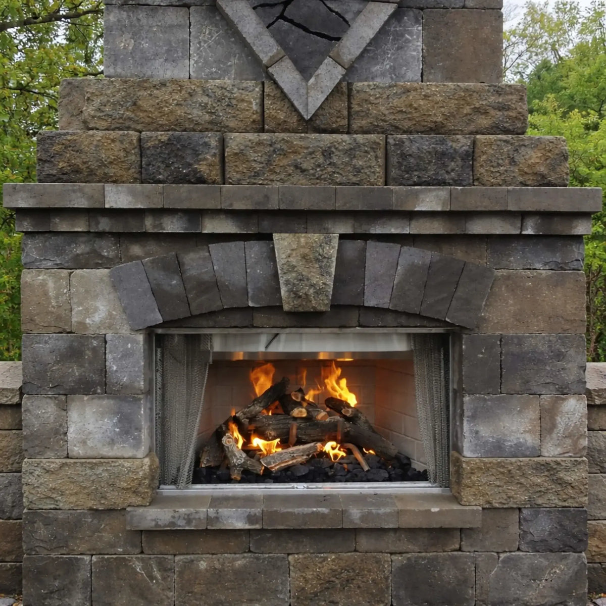 Outdoor stone fireplace with Arizona weathered oak gas logs burning inside a rectangular firebox. The stacked stone surround and arched opening frame glowing flames and realistic logs, creating a warm focal point for an outdoor living area surrounded by greenery.