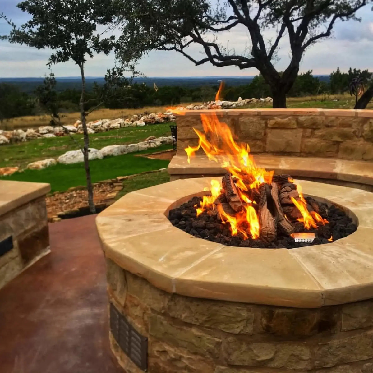 Circular stone fire pit with weathered oak gas logs burning over black lava rock on a scenic patio overlooking rolling hills. The natural stone seating wall and surrounding landscape emphasize a peaceful, elevated outdoor space designed for relaxation and gathering.