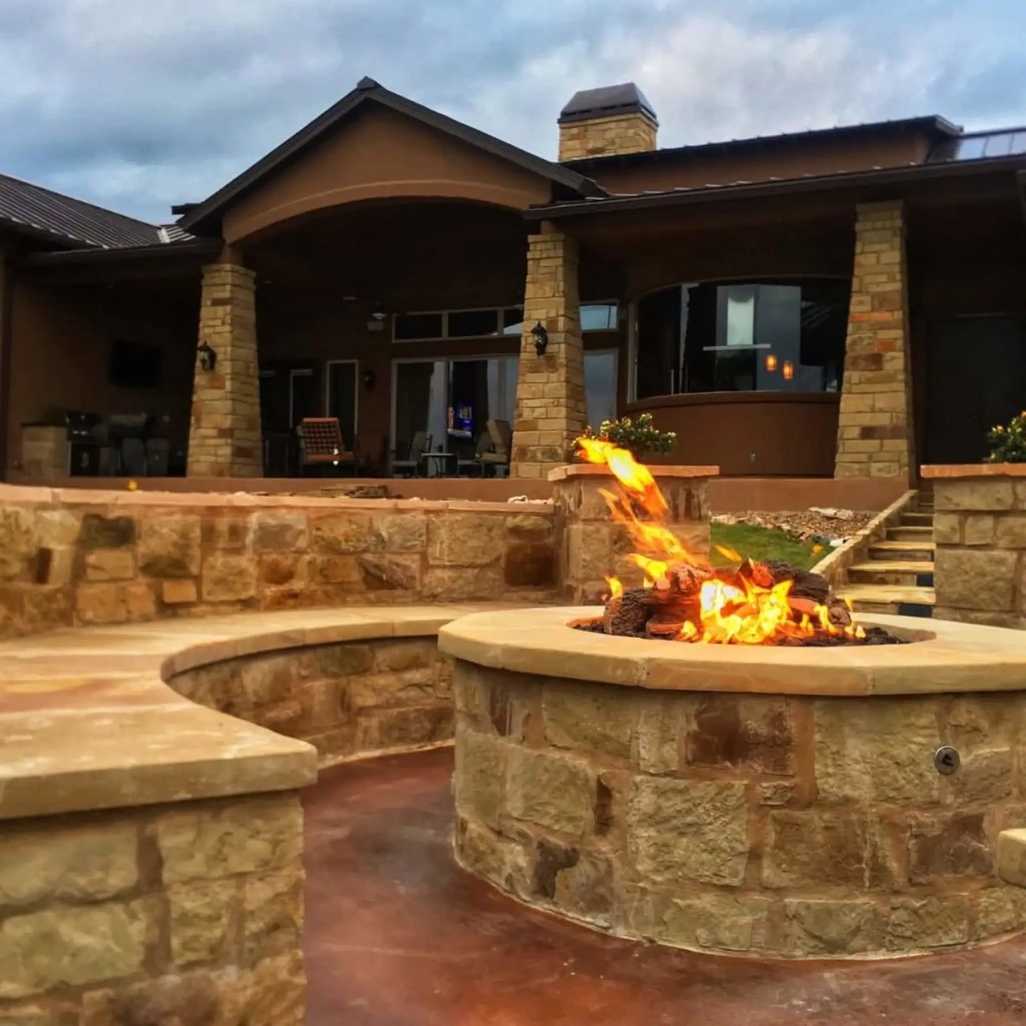 Luxury backyard patio with a round stone fire pit featuring Arizona weathered oak logs and vibrant flames. Positioned near a large home with covered outdoor living space, the fire feature enhances the upscale design with warm ambiance and natural stone finishes.