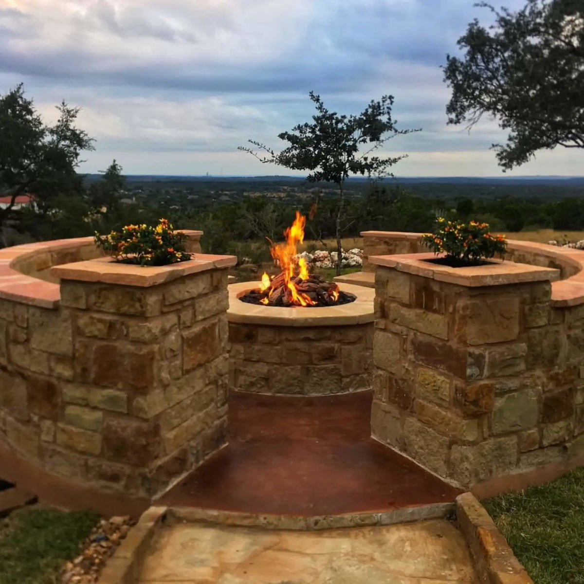 Custom stone fire pit centered between curved seating walls and decorative planters, showcasing weathered oak gas logs with active flames. The elevated patio overlooks a wide landscape, blending natural stone textures with a refined outdoor gathering area.