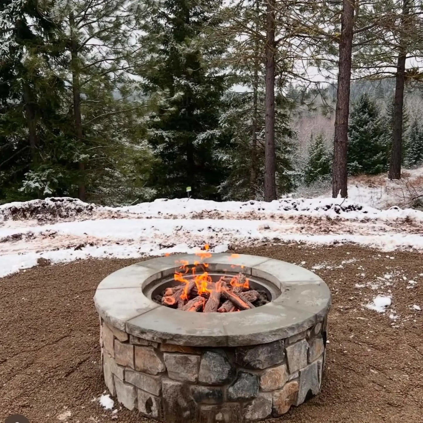 Round stone gas fire pit with weathered oak logs burning in a snowy forest setting. Surrounded by evergreen trees and light snow coverage, the glowing flames contrast against the cold environment, creating a warm and inviting winter outdoor fire feature.